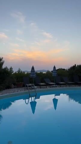 The video shows a swimming pool at sunset. The sky is a light blue with streaks of pink and orange clouds. There are several lounge chairs and umbrellas around the pool, and reflections of them can be seen in the water. In the background, there are trees and a body of water with a distant shoreline. The scene is peaceful and serene.