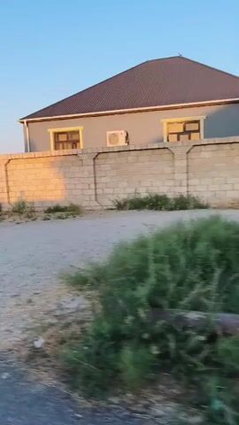 The video begins by showing a house with a gray wall and a brown roof in the background. A stone wall is in the foreground with a patch of grass and dirt in front of it. The camera then pans to the left, revealing two men sitting down. One man is wearing a white and blue striped shirt and blue jeans, while the other man is wearing a maroon t-shirt and blue jeans. They are both looking down. The camera continues to pan left, showing more of the surroundings, which appear to be undeveloped land with some vegetation and buildings in the distance. The sky is clear and blue. The video appears to be shot during the daytime.
