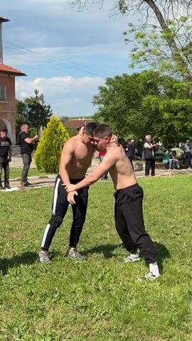 In a wide shot taken during the day, two shirtless young men are grappling in a grassy field, with a small crowd of onlookers surrounding them. The man on the left wears black pants with white stripes down the side and gray sneakers, while the man on the right is in plain black sweatpants and white sneakers. They are locked in a close embrace, each trying to gain an advantage over the other. Behind them, several people stand watching, some taking photos with their phones. A few are seated on a bench in the background. A small yellow shrub stands near the edge of the field, with a building visible in the far background. The sky is partly cloudy.