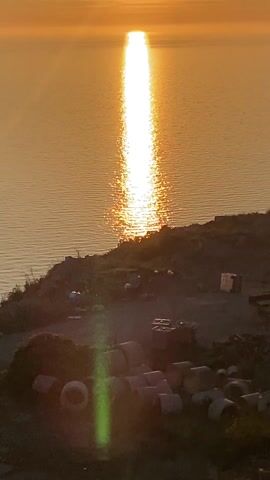 The video shows a daytime shot of the ocean at sunset. The sun is reflecting off the water, creating a bright line down the middle of the image. The foreground shows a small, grassy hill with some debris and pipes scattered around.  The sky is hazy with a few wispy clouds.  A green lens flare appears in the image.