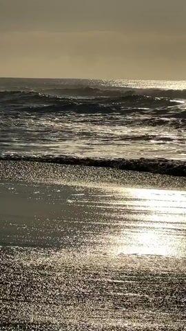A video of a shoreline at sunset shows waves crashing on the beach. The sun is setting and casting a golden glow on the water and sand. The sky is cloudy and the waves are choppy. The video is in landscape mode.