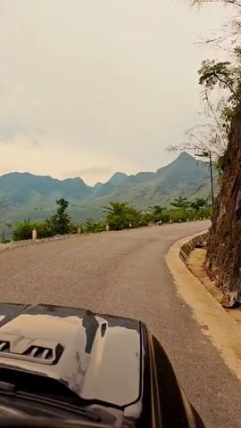 The video depicts a vehicle, likely a car, driving on a paved road winding through a mountainous region. The sky is overcast, casting a muted light over the scene. The road curves gently, lined on one side with lush green vegetation clinging to a rocky cliff face. The other side offers a vista of rolling hills and distant peaks, sparsely dotted with trees. The car's hood is visible in the foreground, providing a driver's perspective of the journey. The overall impression is one of a scenic drive through a natural, somewhat remote landscape.