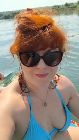 The video begins with a close-up of a woman with red hair and sunglasses wearing a blue bikini top. She is on a paddleboard in a body of water. A person behind her, wearing a black baseball cap and black swim trunks, is paddling with a black paddle. The paddle enters and exits the water, creating ripples. The woman smiles at the camera. The background shows a shoreline with vegetation. The scene appears to be a sunny day.
