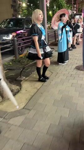 The video shows a street scene at night. In the foreground, two individuals are visible from the waist down. The person on the left is wearing a black outfit with a blue accent and black shoes. The person on the right is wearing a light blue and white outfit with black platform shoes and black stockings. They are standing on a patterned sidewalk.

In the middle ground, several people are standing in a line. The person closest to the foreground on the right is wearing a light blue and white kimono-like outfit and holding a red umbrella. Further down the line, individuals are dressed in black and white outfits, some resembling maid costumes.

In the background, there are trees and buildings with illuminated signs, indicating a commercial or entertainment district. Pedestrians are walking along the street in the distance. The overall atmosphere suggests a lively urban environment, possibly a popular tourist or entertainment area.