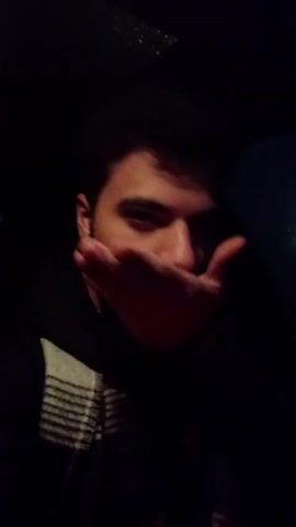 The video begins with a close-up of a young man, looking into the camera with his hand partially obscuring his face. He is wearing a dark hoodie with a striped white and gray pattern on the collar. The camera then shifts to the perspective of the driver of a car, showing a wet road at night. Headlights of oncoming traffic reflect off the wet surface. Streetlights and signs are visible in the background.