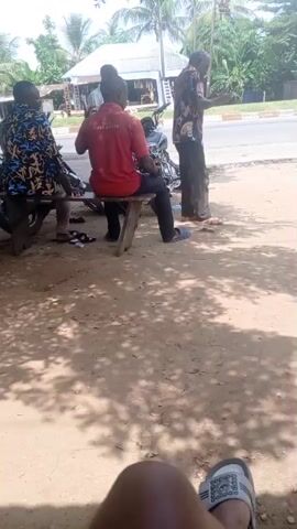 Four men are gathered in a shaded area beside a road. Three of the men are seated on a simple wooden bench. The man on the left wears a blue and gold patterned shirt. The man in the center wears a red shirt with white text. The man on the right is standing, wearing a patterned shirt and dark pants. A motorcycle is parked behind the bench. Across the road, there are buildings, trees, and a red vehicle passes by. The perspective is from someone sitting on the ground, their legs and sandals visible in the foreground. The ground is sandy and covered in leaf shadows.