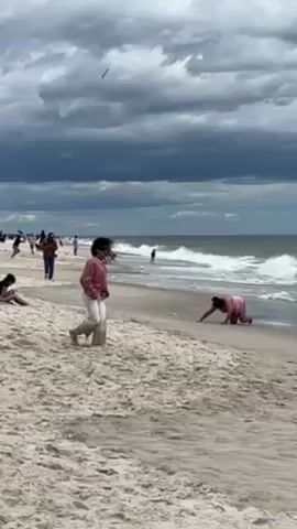 Here is a description of the video:

The video shows a wide, sandy beach on an overcast day. The sky is filled with grey and dark clouds, although some patches of lighter sky are visible towards the horizon. The ocean has waves breaking on the shore.

Several people are scattered along the beach. One person in a pink outfit is kneeling at the water's edge, seemingly playing or digging in the sand. Another person, wearing a pink top and light-colored pants, stands near the center of the frame. Their hair is dark and seems to be blowing in the wind. This person walks slowly along the beach. In the background, other individuals are walking along the shoreline. Some people are wading in the water.

The beach is wide, and the sand is a light beige color. The waves are moderately sized and break with white foam as they reach the shore. The overall impression is one of a cool and windy day at the beach, with people still enjoying the seaside despite the weather.