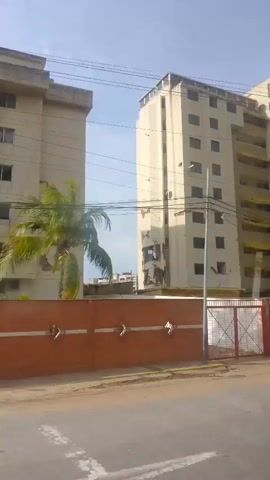 A video shows a view from a vehicle moving past tall, light-colored apartment buildings. The buildings have numerous windows, some with yellow awnings or balconies. One building on the right appears to be under construction or renovation, with visible damage and exposed concrete. A yellow wall with a brick lower section runs along the front of the property, with a metal gate. Power lines are visible overhead. A street with a yellow line is in the foreground, and part of a car is visible at the bottom right. The sky is bright and clear.