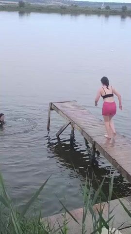 In a wide shot, a young woman in red shorts and a black bikini top is captured mid-jump from a wooden pier into a body of water. Her body is angled, arms outstretched as she plunges downward. To her left, another person, partially submerged in the water, watches her jump. The pier itself is made of weathered wood and is supported by angled wooden beams. The water is calm, with ripples spreading from where the woman is about to land and from the person already in the water. The background features a hazy, distant shoreline with a few trees and what appears to be a power line pylon. The sky is overcast, suggesting a cloudy day, possibly late afternoon or early evening. The foreground is occupied by tall, green reeds or grasses. The overall scene is one of recreational activity, likely a summer day spent swimming and enjoying the outdoors. The facial expressions of the individuals are not clearly discernible, but the activity suggests a playful and possibly daring moment.