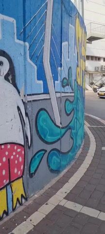 The camera pans along the side of a curved concrete wall covered in graffiti. The bottom portion of the graffiti is blue and green swirling water. Above that is a section that is blue with a green, sea-turtle-like plant and then yellow Hebrew letters. The background shows a sidewalk made of brick and a street with some parked cars.