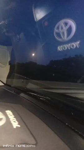The video shows a first-person view from inside a Toyota vehicle at night. The dashboard is visible in the foreground with a Toyota logo embossed on it. Through the windshield, trees and streetlights can be seen, suggesting the vehicle is on a road. The moon is also visible in the sky. The camera appears to be mounted on the dashboard or windshield. The footage is shaky, indicating movement.