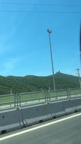 The media file shows a scenic view of a mountainous landscape on a clear day. The foreground features a road with a concrete barrier and a metal fence, suggesting a highway or elevated road. A tall lamppost stands prominently on the right side of the road.

Beyond the road, lush green hills dominate the midground, covered in dense vegetation. A distinctive rock formation, appearing like a single, large boulder perched atop a smaller hill, stands out against the sky. This unique geological feature is a focal point of the scene.

The sky is a vibrant, clear blue, indicating fair weather and likely daytime. The sun appears to be high, casting shadows, though not intensely. The overall impression is one of natural beauty and a sense of travel or transit.

The scene is set in Hoa Xuan, Vietnam. The presence of the road infrastructure and the tropical vegetation are consistent with this location. There is no visible text in the media file. The emotion evoked is one of serenity and appreciation for the natural landscape.