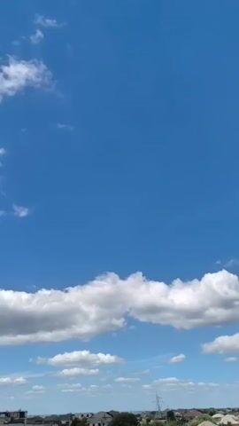 The video is a vertical shot of a bright blue sky with scattered white clouds. The bottom portion of the frame shows the rooftops of houses in a rural or suburban setting, with some trees and fields visible in the distance. The camera appears to be looking slightly downwards from a high vantage point.
