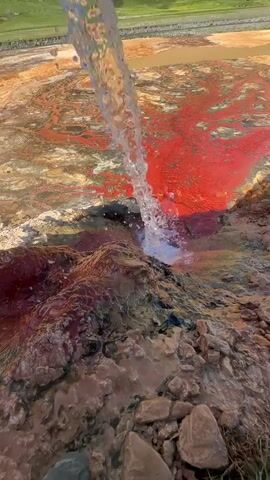 This media file captures a vibrant natural phenomenon in Ketrisi, Georgia. A strong stream of clear water is cascading down from an unseen source above, hitting a rocky, reddish-brown mineral deposit. The impact of the water creates a splash and sends droplets flying. The water then flows into a shallow pool characterized by striking red and orange mineral deposits, creating a colorful, almost abstract landscape. The surrounding terrain is rugged and rocky at the foreground, transitioning to a barren, mineral-rich plain, and finally to rolling green hills in the background under a bright, blue sky with scattered clouds, suggesting a clear, sunny day. The scene is devoid of people. The overall impression is one of raw, natural beauty and geological activity, with the vibrant colors of the mineral deposits being a prominent feature. There is no visible text in the media file.