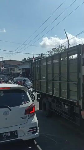 The video shows a busy street scene with a lot of traffic. There are several cars and motorcycles on the road. People are riding on motorcycles, and some are walking. The buildings on the side of the street appear to be shops. The sky is clear and blue. There are power lines running across the street. The overall impression is one of a crowded and active urban environment.