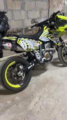 A yellow and black supermoto motorcycle is parked inside a dimly lit, industrial-looking space with brick and concrete walls. A person, partially obscured and wearing dark clothing, is leaning over the motorcycle. Another person, also in dark clothing, is standing further back in the space, near some equipment. The space appears to be a workshop or garage, with a concrete floor and exposed brickwork. The lighting is low, casting shadows on the floor and walls.