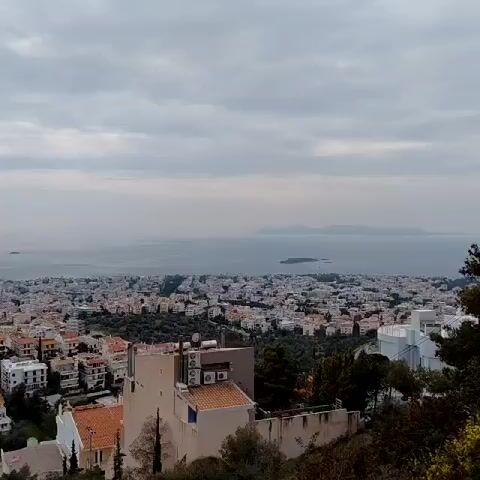 The video shows a high-angle view of a city stretching towards the ocean under a cloudy sky. The city is densely populated with buildings, mostly light-colored with terracotta roofs.  The ocean is visible in the distance, with islands dotting the horizon. The foreground includes trees and the rooftops of buildings closer to the camera's vantage point.