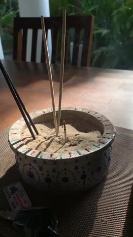 This media file shows a close-up shot of an incense holder filled with sand, placed on a wooden table. Three incense sticks are burning, emitting thin trails of smoke. Two of the incense sticks are standing upright in the center of the holder, while a third, darker incense stick is positioned to the left and leaning. The incense holder itself is round and decorated with colorful circular patterns.

In the foreground, a set of keys and a small rectangular tag are visible. The tag appears to be from a business, with text that reads "TVS WASHANTHA MOTORS Galle" and a phone number.

The background suggests an indoor setting with a glimpse of outdoor greenery through a window. A wooden chair with vertical slats is also visible, partially out of focus. The lighting suggests it is daytime, with sunlight casting shadows. The overall mood is peaceful and serene, possibly indicating a moment of meditation or relaxation. The location context is identified as Kathaluwa, Sri Lanka.