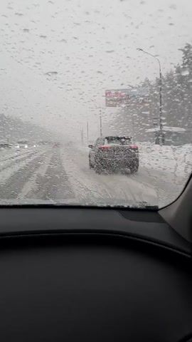 The view from inside a car driving on a snowy road. A dark-colored car drives ahead. A billboard is seen to the right. The windshield is covered in water droplets. Snow is piled on either side of the road. Streetlights line the road. Other vehicles are visible in the distance. It is snowing. Music is playing.