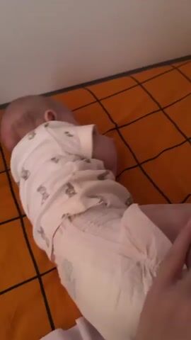 The video shows a baby lying on their stomach on an orange and black plaid surface, likely a blanket or sheet on a bed. The baby is wearing a white short-sleeved onesie with a ribbed texture and a pattern of small bunnies. A white diaper is visible on the baby's lower half. An adult hand, with light pink nail polish, is gently patting or holding the baby's diaper area from the side. The baby's head is turned away from the camera, and they appear to be resting or sleeping peacefully. Part of the text "joories" and "Marimelo" is visible on the diaper. The lighting suggests an indoor setting.