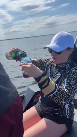 A woman wearing a white baseball cap, sunglasses, and a black and gold fishing shirt is on a boat. She is pouring liquid from a green bottle into a clear plastic cup. The cup already contains a red liquid. The boat is on the water, and the sky is blue with some clouds. There is another person partially visible on the left side of the frame. Fishing rods are visible in the background.