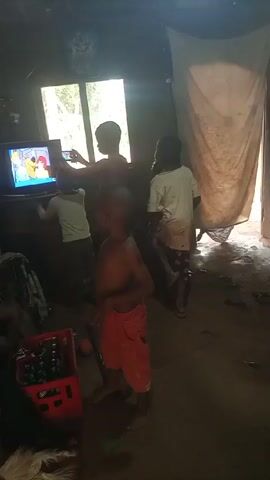 The video shows a dark room with a dirt floor and a window. Four children are gathered around a television that is playing a cartoon. One child is holding up a phone and appearing to film the TV screen. There is a red crate full of bottled drinks and some other miscellaneous items on the floor. A light-colored curtain hangs in the background.