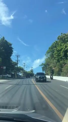 The video captures a forward-facing view from inside a vehicle driving on an asphalt road under a clear blue sky with scattered clouds. The road has yellow lane markings. Several other vehicles are visible: a black pickup truck in the right lane, a red car in the distance, and a white SUV ahead of the red car. Trees and foliage line both sides of the road, with numerous utility poles and overhead wires present on the left. The sun casts long shadows across the road, indicating it is likely daytime. The overall environment appears to be a tropical or sub-tropical area due to the lush greenery.