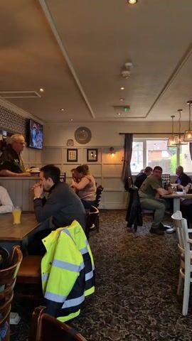 The camera pans through the interior of a pub, with several patrons seated at tables, eating and drinking. A man wearing glasses and a black jacket is standing near a group of people, while a high-visibility jacket hangs on a chair. A large TV screen displays a soccer team celebration, with the text "FA Cup Winners 2023" visible. Other objects visible include chairs, tables, pendant lights, wall decorations, and various signs. The pub has a patterned carpet and wood paneling.