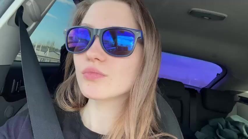 A young woman is inside a car, wearing sunglasses and a seatbelt. She is looking toward the camera, her expression neutral. The car appears to be moving, with outside scenery visible through the windows. There is a black object on the back seat. The car's interior is visible.