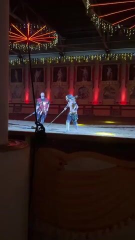 Two men in costume are sword fighting in an arena. One man, wearing a red and gold tunic, is on the left, holding a sword and shield. The other man, wearing dark pants and a gold belt, is on the right, holding a sword. The arena is decorated with Roman-style murals and red lighting. The floor is covered in sand.