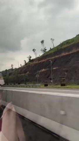 The video captures a view from a vehicle traveling on a highway. On the right side of the road, there is a steep, rocky embankment with some sparse vegetation and a few trees. The embankment appears to have been reinforced with some form of netting or material. The sky is overcast with heavy, grey clouds. A white truck is visible on the highway in the distance, traveling in the same direction as the camera vehicle. The foreground shows a concrete barrier separating the lanes of traffic, with the reflection of the interior of the vehicle visible on its surface. The road appears wet, suggesting it may have recently rained or is still raining lightly. The overall environment is a highway passing through a hilly, vegetated area under a cloudy sky.