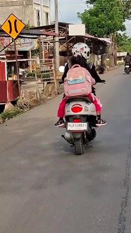 A person wearing a black jacket and a white helmet with black spots is riding a silver scooter. A young girl wearing a pink jacket, black pants, and pink shoes is riding behind the person, facing forward and wearing a pink backpack. The scooter is moving on a paved road, with buildings and vegetation visible in the background. The license plate of the scooter reads "BL 6148 MAV." The video is shot from behind the scooter, following it as it moves away from the camera.
