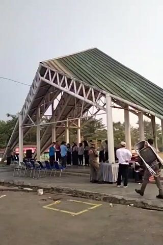 A group of people, some wearing uniforms, are gathered under a covered structure with a green, sloped roof supported by white columns. Several chairs are set up in front of the structure. Some individuals are standing around tables draped with cloths, while others are sitting. One person is carrying a folded chair. The scene appears to be an outdoor event or gathering, possibly a ceremony or meeting.