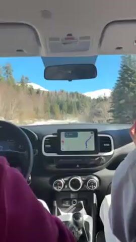 Here's a description of the video:

The video is shot from the interior of a moving car, looking out through the windshield.  Two people are visible in the front seats, one wearing a pink/purple shirt and one wearing white. The dashboard is visible, including the steering wheel, instrument panel, infotainment screen showing a GPS navigation, and the climate control panel.

Outside the car, the scene is a road winding through a wintery landscape. There are trees on either side of the road, and snow on the ground in patches. In the distance, snow-capped mountains are visible against a clear blue sky.

Music is playing in the background.