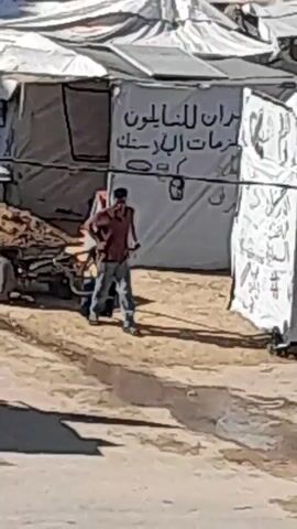 The video depicts a scene in what appears to be a camp or settlement with temporary structures made of white fabric. In the foreground, a man in a red shirt and blue jeans is walking towards the right. To his left, a two-wheeled cart loaded with what looks like dirt or debris is being pushed by a person wearing a white hat and shirt. Another person, possibly a child, walks across the foreground from left to right. In the background, the fabric structures have signs with Arabic writing. The ground is sandy and uneven. The overall impression is one of a working environment within a makeshift settlement.