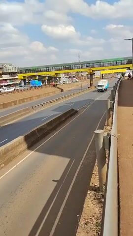 The video, shot from an elevated pedestrian walkway, presents a view of a multi-lane highway under a partly cloudy sky. The highway is divided by concrete barriers. A white and blue truck travels along the highway from right to left.  In the background, on the other side of the highway, are buildings, parked cars, and a yellow pedestrian bridge. Towards the end of the video, a man in a white shirt and dark pants walks along the pedestrian walkway to the right of the camera.