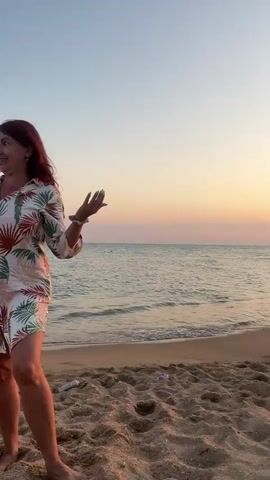 The video captures a sunset scene at a beach. The sky transitions from a pale blue at the top to a vibrant orange and pink closer to the horizon. The sun, a bright white orb, is visible low in the sky, casting a golden reflection on the water. Gentle waves lap the sandy shore. In the distance, a small white boat is visible on the water. Several people are scattered along the beach, some wading in the water and others walking along the shoreline. A few pieces of debris, including what appears to be driftwood, are present on the sand. The sand itself has ripples and patterns from the wind and waves. The overall impression is a serene and picturesque evening at the beach.