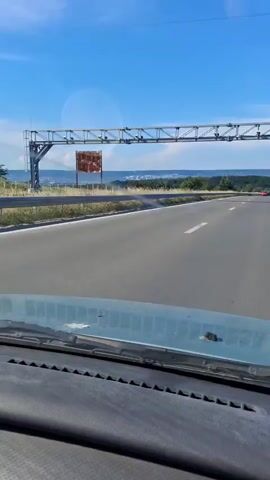 The video is captured from the interior of a moving vehicle, looking forward through the windshield. The perspective is from the driver's seat, with the dashboard and windshield wipers visible in the foreground. The road ahead is paved and appears to be a highway or a similar multi-lane road, with a white dashed line marking the lanes. To the left of the road, a metal guardrail runs along the edge, with dry grass and some vegetation behind it. Further beyond the guardrail, a large metallic structure, possibly an overhead signpost, is visible. In the distance, a rolling landscape with trees and a body of water can be seen under a clear blue sky with scattered clouds. The lighting suggests it is daytime, likely sunny. The camera is static within the moving vehicle. There are no people visible in the frame. The overall scene depicts a journey on a road through a scenic, possibly rural or semi-rural, environment.