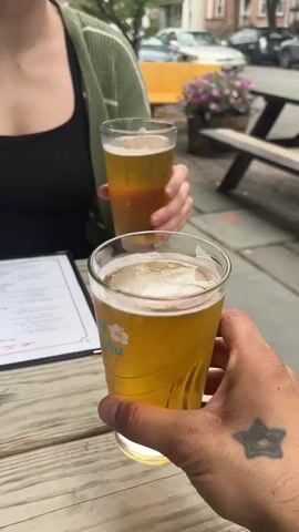 The video shows two people at an outdoor table, drinking beer. A hand with a star tattoo holds up a glass of light-colored beer, embossed with a flower design. Behind it, a woman in a green cardigan and black tank top is holding another glass of beer. The camera moves to show her taking a sip of the beer. There is a menu on the table in front of her. Other people are visible in the background, along with cars and buildings. The table is made of wood.