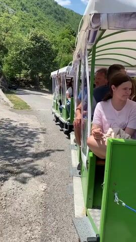 A green train with a white canopy transports several passengers through a mountainous, wooded area on a sunny day. The train is moving along a gravel and asphalt path. On the left side of the path, there is grass and rocks. In the background, there are more trees and mountains. Two white cars and a small building are visible in the distance.