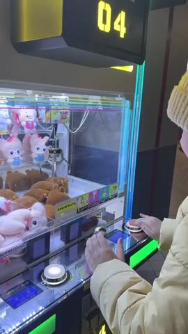 The video shows a young girl playing a crane machine in an indoor entertainment venue. The machine is brightly lit with multicolored lights and has a digital display showing the number "04". Inside the machine, a variety of plush toys, including bears, cats, and pigs, are visible. The girl, wearing a yellow jacket and a white beanie, operates the machine using a joystick and buttons. She watches intently as the crane moves, showing anticipation and surprise as it picks up a plush toy. The background suggests a shopping mall or arcade setting.