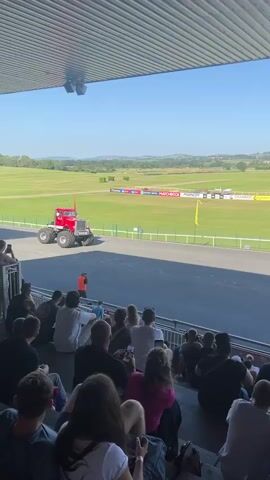 A large red monster truck with oversized tires is positioned on a paved track. Spectators are seated in bleachers overlooking the track. Behind the truck and track, a green field stretches out towards a distant treeline under a clear blue sky. The monster truck is at the start of a ramp structure. Two people are visible standing near the truck. The truck then drives onto the ramp and crushes several cars parked at the bottom.