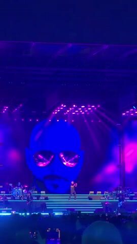 The video shows a concert with a large blue projection of a man's face wearing sunglasses on the stage backdrop. A singer is performing on a lit-up stage in front of the projection, with a band playing behind him. The stage is lit with purple and pink lights, and there are spotlights in the background. A crowd of people is visible in the foreground, holding up their phones to record. The atmosphere is energetic and exciting.
