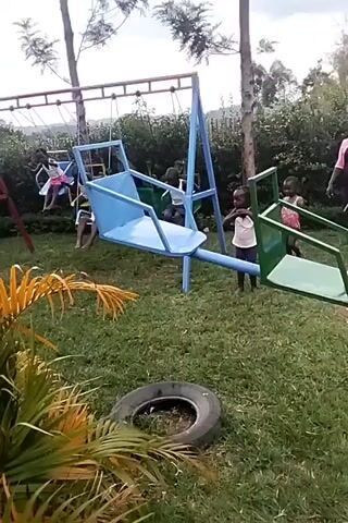The video shows a small playground with children. There are three swingsets with children sitting on the swings. A blue see-saw type structure has a child in one seat, while a young child in a pink shirt stands nearby. A green see-saw is to the right with another child standing near it. The ground is grass, and a tire lies near the bottom left of the frame.  There are trees in the background.