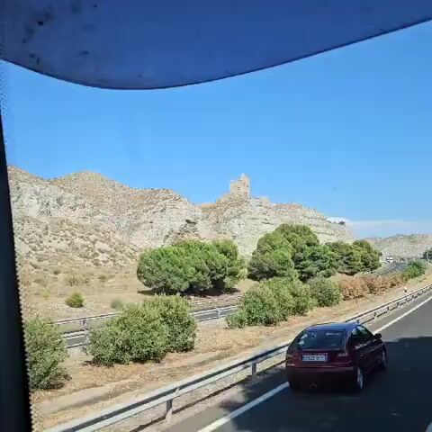 A video of a crumbling castle ruin atop a rocky hill. The castle is in disrepair with visible holes and missing sections. The hill is covered in sparse vegetation and some trees are visible in the foreground. The sky is clear and blue.