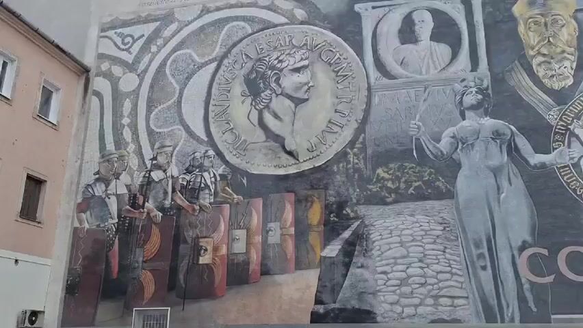 The video shows a large mural painted on the side of a building. The mural depicts various historical and symbolic images, including Roman soldiers with shields, a large coin with a Roman emperor's profile, a statue of a woman holding a staff, portraits, and scenes of cityscapes. The mural appears to be designed in a collage style, combining elements from different eras and sources. The adjacent building is painted in light pink, with white framed windows. The overall impression is that the mural tells a story or commemorates the history of a specific place.