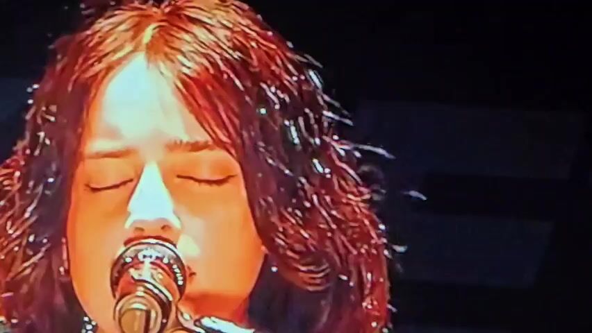 The media file shows a close-up of a young woman with reddish-brown, curly hair, singing into a microphone on a dimly lit stage. Her eyes are closed, and her mouth is open as she sings. She is wearing a light-colored shirt with dark stripes. The background is dark, with some faint lights visible, suggesting a performance or concert setting. The overall mood appears to be intimate and emotional, likely captured during a musical performance. There is no visible text in the media file. The location context provided is Satbayev, Kazakhstan.