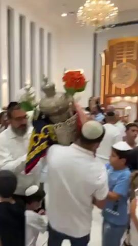 The video shows a crowd of people, mostly men and boys wearing yarmulkes, moving through a brightly lit room with white walls and large windows. Several men are carrying ornately decorated objects, possibly Torah scrolls, adorned with flowers and fabric. People are talking, singing, and seem to be celebrating. The setting appears to be a synagogue or religious gathering space with a prominent chandelier and decorative woodwork.