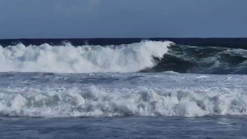 The video shows ocean waves crashing towards the shore. The waves are white and foamy, with dark blue water underneath. The horizon is visible in the distance. The sky is a dull blue. There are no people visible in the video.
