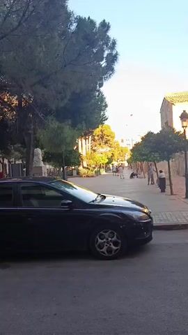 The video shows a street scene in what appears to be a European town. Several cars are parked along the street, and people are seen walking and sitting in a small park or plaza in the distance. The buildings have a classic, old-world style architecture. The sky is blue. The camera pans slowly to the right, revealing more of the scene. Several trees are visible, some of which appear to be bare. Overall, the scene appears calm and peaceful.